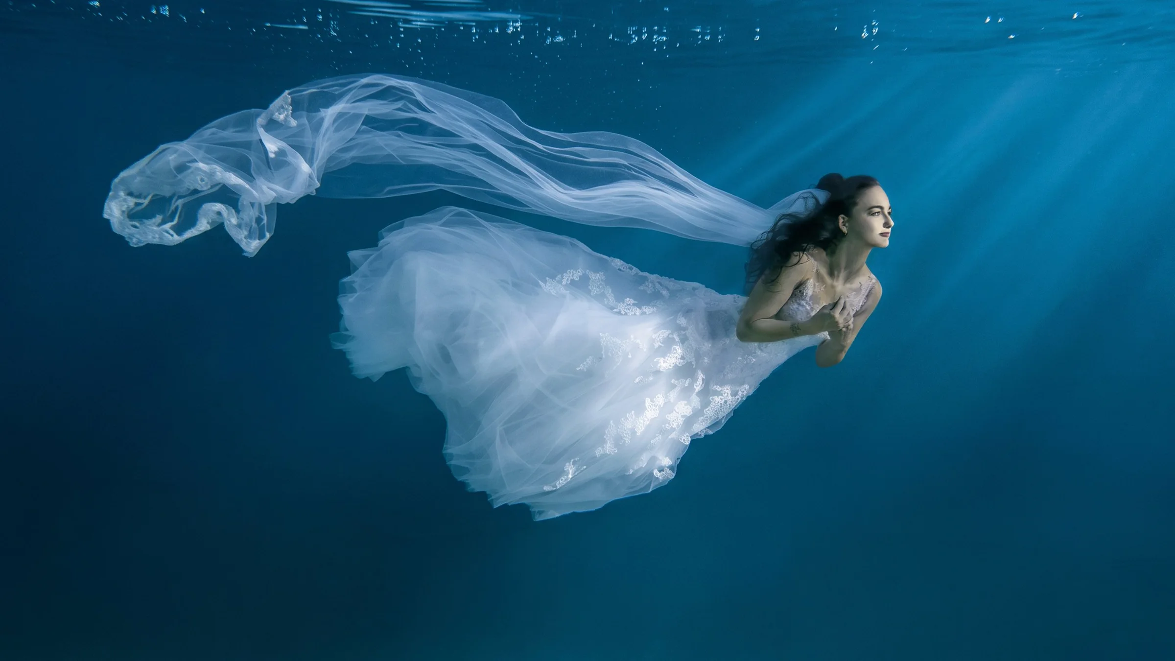 Braut im weissen Spitzenkleid mit langem Schleier schwebt im Lichtstrahl unter Wasser