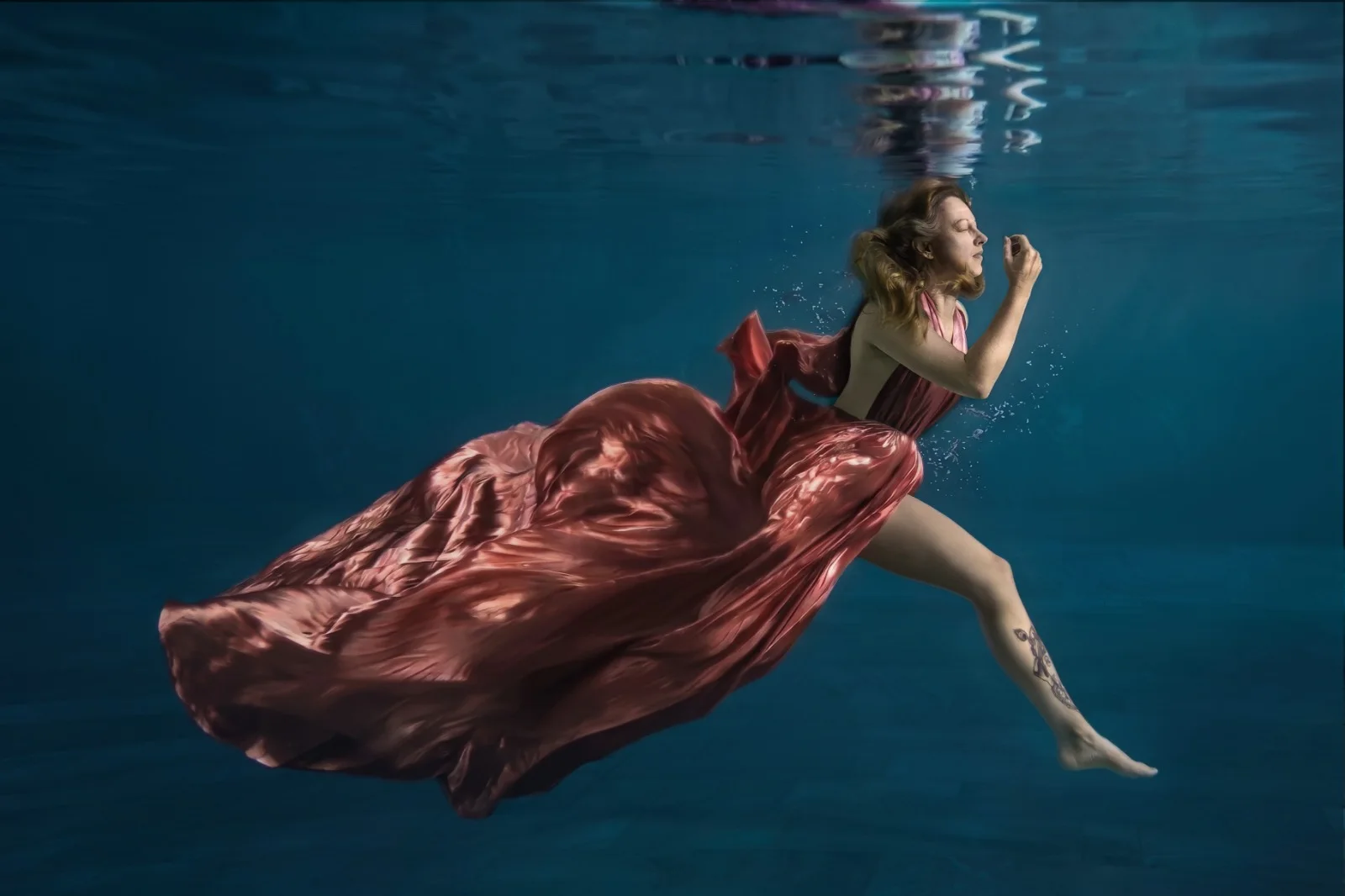 Frau im fliessenden roséroten Satinkleid gleitet mit geschlossenen Augen durch tiefblaues Wasser – Beyond Gravity