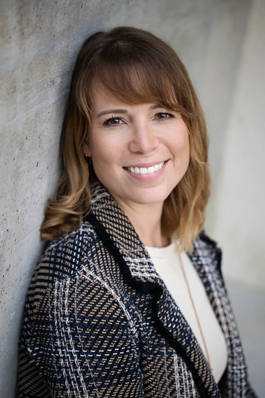 Frau im Tweed-Blazer lehnt lachend an einer Betonwand, Portrait mit Struktur im Stoff