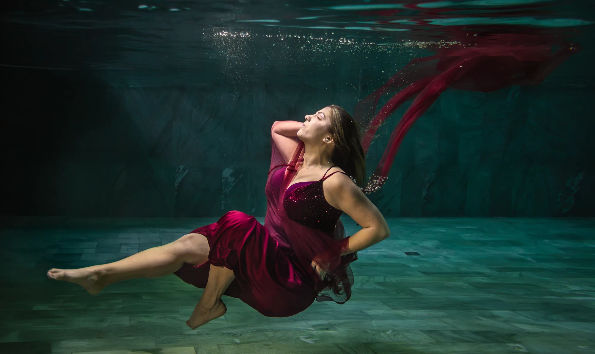 Frau im weinroten Samtkleid schwebt mit geschlossenen Augen unter Wasser, rote Tücher treiben zur Oberfläche – Unterwasser-Fotoshooting